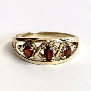 May include: A gold ring featuring three oval-cut dark red garnet gemstones set in a horizontal row. The band has a polished gold finish with an openwork design that frames each of the three stones, creating a continuous, interconnected pattern.