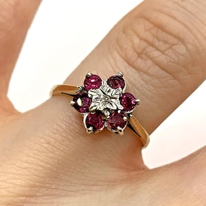 May include: A gold ring featuring a flower-shaped cluster of six round, deep pink gemstones surrounding a central silver-toned metal element with a small, clear, round stone set in the middle of the floral arrangement.