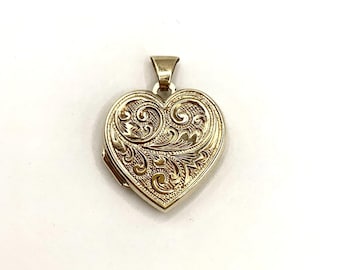 May include: A gold-toned heart-shaped locket pendant featuring an intricate, embossed scrollwork pattern across the front surface. The pendant includes a small, polished metal bail at the top for attachment to a chain or necklace cord.