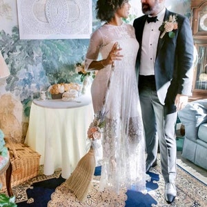 May include: A couple in a wedding ceremony, the bride is holding a broom decorated with flowers, the groom is wearing a black tuxedo, the bride is wearing a white lace dress, the ceremony is taking place in a room with a floral wallpaper and a table with a white tablecloth.