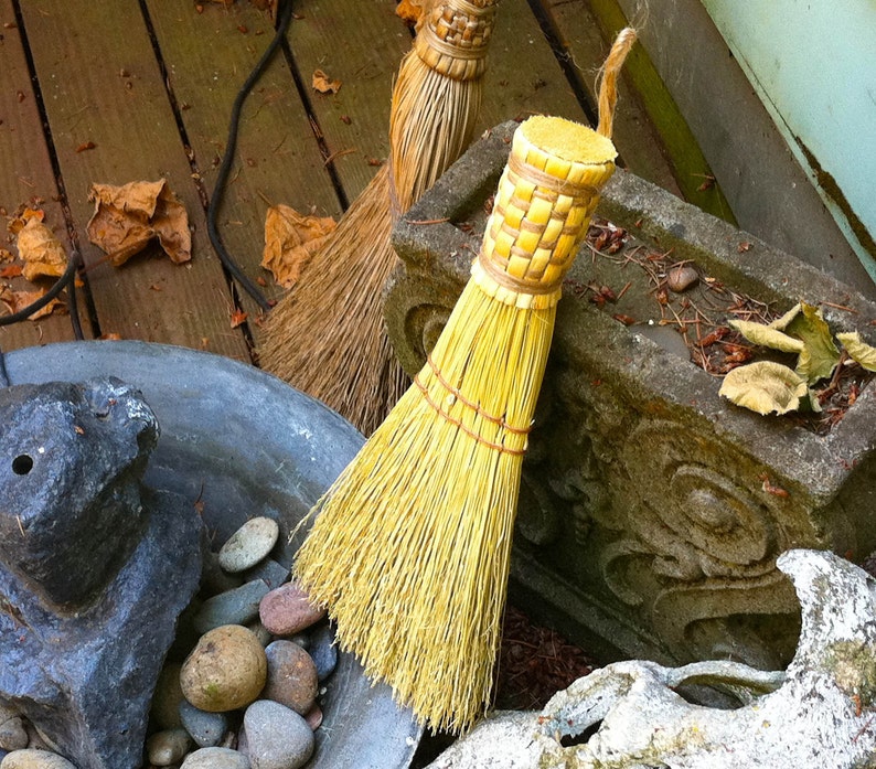 Pu&ograve; includere: Due scope fatte a mano con setole di paglia naturale. Una scopa ha un manico marrone chiaro e l'altra un manico giallo. Le scope sono su una superficie di legno.