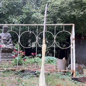 May include: A white broom with a long wooden handle leaning against a white metal fence. The broom is in front of a green bush and a statue of a person sitting on a stone base.