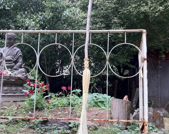 May include: A white broom with a long wooden handle leaning against a white metal fence. The broom is in front of a green bush and a statue of a person sitting on a stone base.