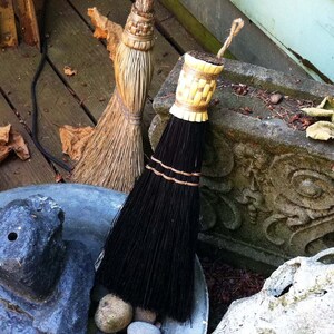 Pu&ograve; includere: Due scope con setole naturali, una con un manico nero e l'altra con un manico marrone, sono appoggiate a un muro di pietra. La scopa con il manico nero ha una fascia di paglia intrecciata intorno.
