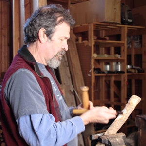 K&ouml;nnte beinhalten: Ein Mann in einem blauen Hemd und einer roten Weste schnitzt mit einem Messer und einem Holzhammer ein St&uuml;ck Holz. Er arbeitet an einem Holzprojekt in einer Werkstatt.