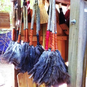 May include: A collection of handmade feather dusters with natural wooden handles. The dusters feature dark gray feathers and are wrapped with leather in various colors, including brown, red, and black. The dusters are hanging on display.