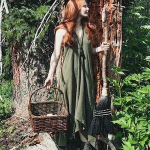 Peut inclure: Une femme en robe verte se tient dans une for&ecirc;t, tenant un panier tiss&eacute; et un balai noir. Elle porte des bottes noires et un collier de perles.