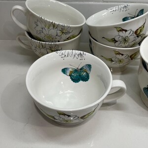 Puede incluir: Tazas de t&eacute; de cer&aacute;mica blanca con asas, decoradas con dise&ntilde;os florales y de mariposas en verde, amarillo y turquesa. Las tazas est&aacute;n apiladas y dispuestas sobre una superficie blanca, mostrando la delicada obra de arte.
