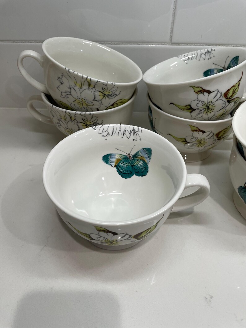 Puede incluir: Tazas de t&eacute; de cer&aacute;mica blanca con asas, decoradas con dise&ntilde;os florales y de mariposas en verde, amarillo y turquesa. Las tazas est&aacute;n apiladas y dispuestas sobre una superficie blanca, mostrando la delicada obra de arte.