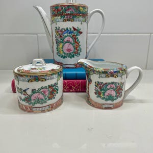 May include: A porcelain tea set consisting of a tall teapot with a lid, a lidded sugar bowl, and a creamer. Each piece features a white base with colorful hand-painted floral motifs, butterflies, and gold trim accents in a repeating pattern.