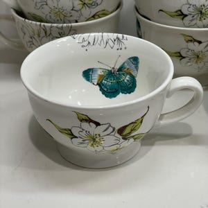 Puede incluir: Tazas de t&eacute; de cer&aacute;mica blanca con un dise&ntilde;o floral y de mariposas. Las tazas tienen un asa y una abertura ancha. El interior de una taza presenta una mariposa turquesa y amarilla. El exterior est&aacute; decorado con flores blancas y hojas verdes.