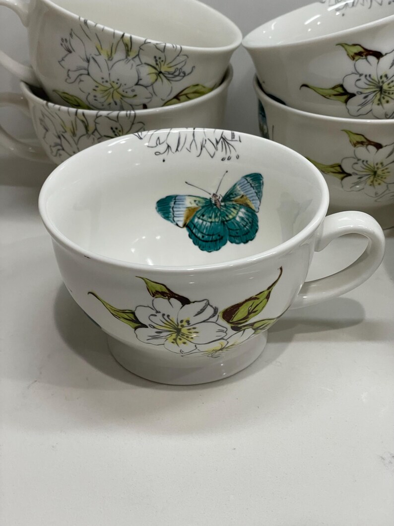 Puede incluir: Tazas de t&eacute; de cer&aacute;mica blanca con un dise&ntilde;o floral y de mariposas. Las tazas tienen un asa y una abertura ancha. El interior de una taza presenta una mariposa turquesa y amarilla. El exterior est&aacute; decorado con flores blancas y hojas verdes.