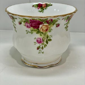 May include: A white ceramic vase with a scalloped rim and base, adorned with a floral rose pattern in shades of pink, red, and yellow. The vase has a gold trim and a rounded shape, suitable for displaying flowers.
