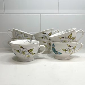 Puede incluir: Un juego de siete tazas de t&eacute; de cer&aacute;mica blanca con asas. Cada taza est&aacute; decorada con un dise&ntilde;o floral en amarillo, verde y azul. Una taza presenta una mariposa azul. Las tazas est&aacute;n dispuestas sobre una superficie blanca.