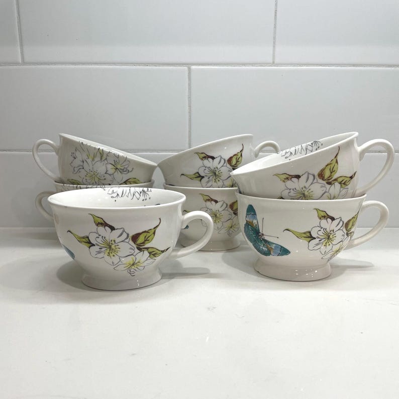 Puede incluir: Un juego de siete tazas de t&eacute; de cer&aacute;mica blanca con asas. Cada taza est&aacute; decorada con un dise&ntilde;o floral en amarillo, verde y azul. Una taza presenta una mariposa azul. Las tazas est&aacute;n dispuestas sobre una superficie blanca.
