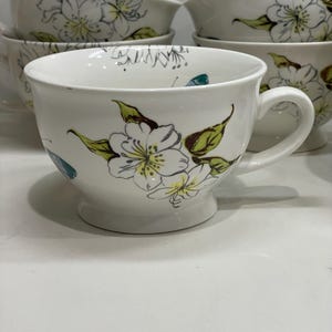 Puede incluir: Tazas de t&eacute; de cer&aacute;mica blanca con un dise&ntilde;o floral. Las tazas tienen una forma ancha y redondeada con un asa y una base de pedestal. El dise&ntilde;o floral presenta flores blancas con centros amarillos y hojas verdes. Tazas adicionales apiladas en el fondo.