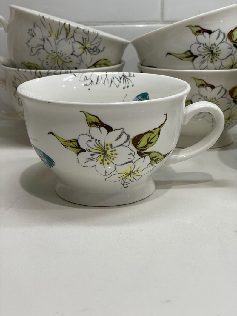 Puede incluir: Tazas de t&eacute; de cer&aacute;mica blanca con un dise&ntilde;o floral. Las tazas tienen una forma ancha y redondeada con un asa y una base de pedestal. El dise&ntilde;o floral presenta flores blancas con centros amarillos y hojas verdes. Tazas adicionales apiladas en el fondo.