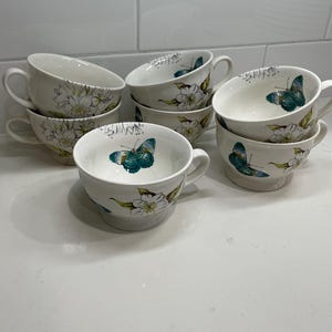 Puede incluir: Un conjunto de tazas de t&eacute; de cer&aacute;mica blanca con asas. Cada taza est&aacute; decorada con un dise&ntilde;o floral y de mariposas en tonos verdes, azules y amarillos. Las tazas est&aacute;n apiladas y dispuestas sobre una superficie blanca.