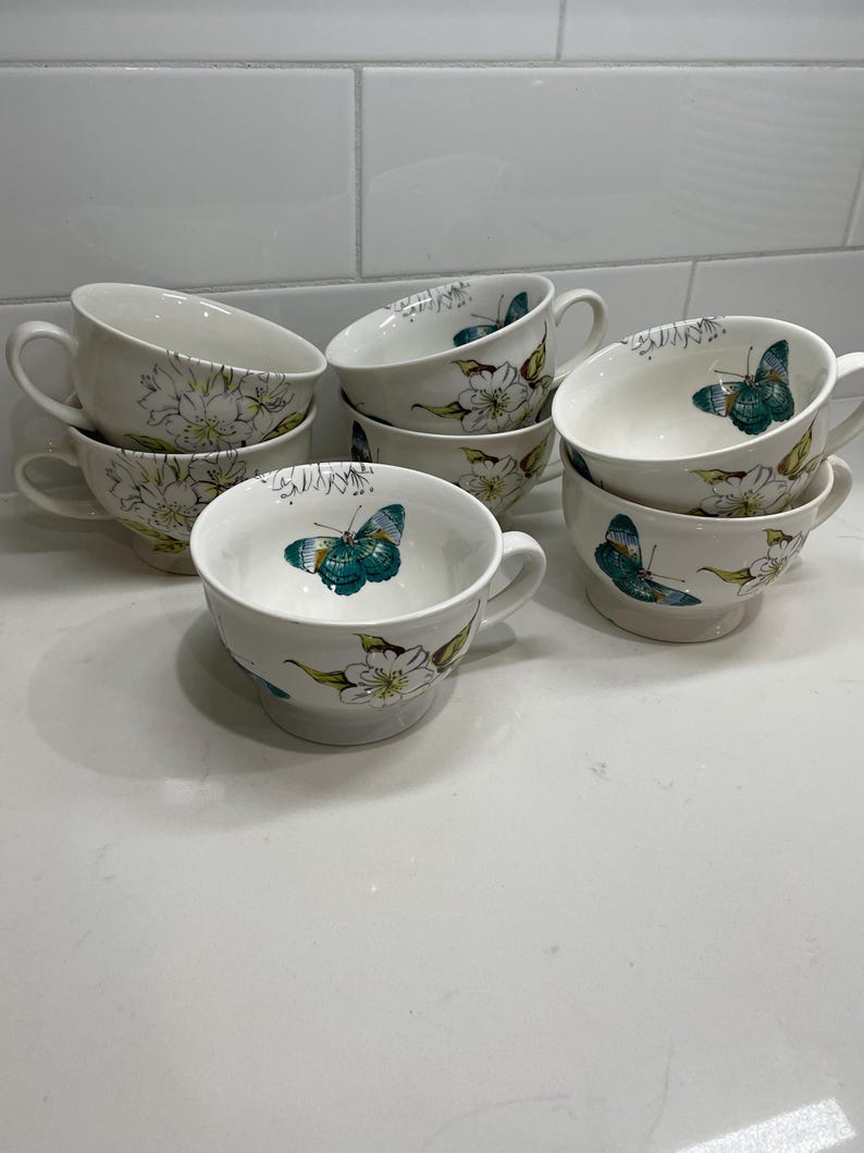 Puede incluir: Un conjunto de tazas de t&eacute; de cer&aacute;mica blanca con asas. Cada taza est&aacute; decorada con un dise&ntilde;o floral y de mariposas en tonos verdes, azules y amarillos. Las tazas est&aacute;n apiladas y dispuestas sobre una superficie blanca.