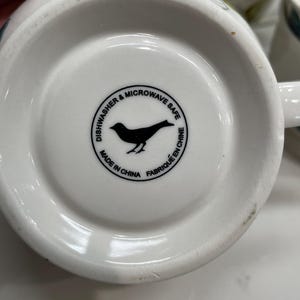 Puede incluir: Base de taza de cer&aacute;mica blanca con una silueta de p&aacute;jaro negro dentro de un c&iacute;rculo que dice "Dishwasher & Microwave Safe" y "Made in China Fabriqu&eacute; en Chine".