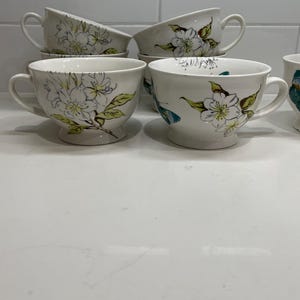 Puede incluir: Tazas de t&eacute; de cer&aacute;mica blanca con platillos a juego. Cada taza presenta un dise&ntilde;o floral con flores blancas, hojas verdes y detalles amarillos. Las tazas tienen forma redondeada y base de pedestal. Las asas son curvas. Los platillos son redondos.