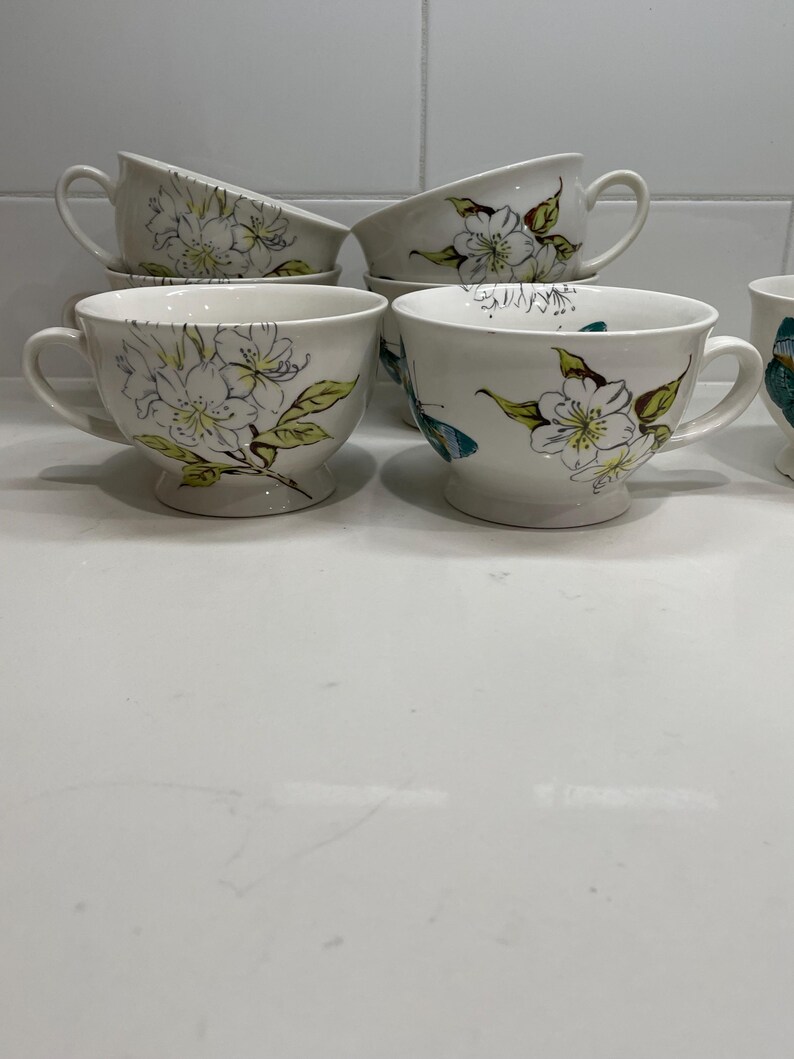 Puede incluir: Tazas de t&eacute; de cer&aacute;mica blanca con platillos a juego. Cada taza presenta un dise&ntilde;o floral con flores blancas, hojas verdes y detalles amarillos. Las tazas tienen forma redondeada y base de pedestal. Las asas son curvas. Los platillos son redondos.