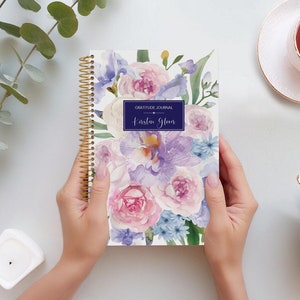 May include: A white spiral bound gratitude journal with a watercolour floral design featuring pink, purple, and blue flowers. The journal cover has the text "Gratitude Journal" and "Karsten Glover" printed in a navy blue rectangle.