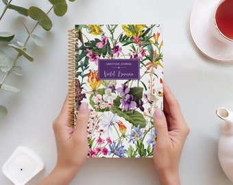 May include: A spiral bound gratitude journal with a floral design featuring purple, pink, yellow, and white flowers. The cover reads "Gratitude Journal Violet Benson".