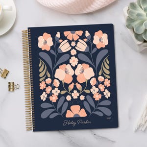 May include: A navy blue spiral-bound planner with a floral design in peach and white. The cover features a stylized flower pattern with the name "Haley Parker" and the year "2023" printed in white. Gold binder clips and a cup of coffee are visible.