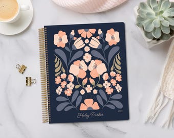 May include: A navy blue spiral-bound planner with a floral design in peach and white. The cover features a stylized flower pattern with the name "Haley Parker" and the year "2023" printed in white. Gold binder clips and a cup of coffee are visible.