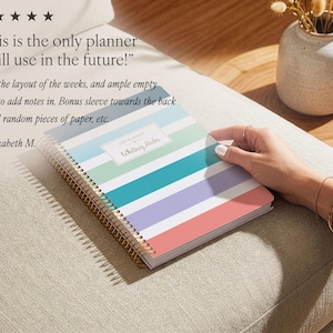Puede incluir: Una agenda de espiral con rayas horizontales en tonos azul verdoso, azul claro, lavanda y coral. La portada incluye una etiqueta rectangular blanca con el texto "2023 PLANNER" y "Whitney Hicks" impreso en una fuente cursiva oscura.