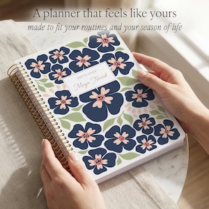 May include: A spiral-bound 2022 planner with a white cover featuring a dark blue and light pink floral pattern and green leaves. A central white rectangular label displays the text 2022 PLANNER and Maya Novak in a script font. The coil is gold-toned.