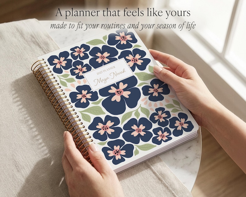 May include: A spiral-bound 2022 planner with a white cover featuring a dark blue and light pink floral pattern and green leaves. A central white rectangular label displays the text 2022 PLANNER and Maya Novak in a script font. The coil is gold-toned.