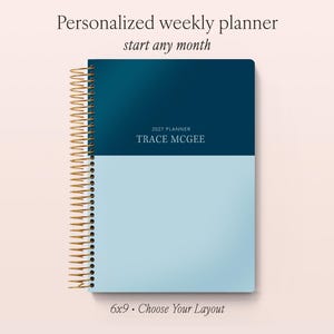 May include: A personalised weekly planner with a two-tone cover featuring a dark blue top half and a light blue bottom half. The cover displays the text 2027 PLANNER TRACE MCGEE. It has a gold-coloured metal spiral binding on the left side.