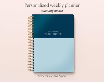 May include: A personalized weekly planner with a two-tone cover featuring a dark blue top half and a light blue bottom half. The cover displays the text 2027 PLANNER TRACE MCGEE. It has a gold-colored metal spiral binding on the left side.