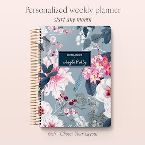 May include: A spiral-bound weekly planner with a blue-grey cover featuring a floral pattern of pink, white, and dark blue flowers and leaves. A central dark blue rectangular label displays the text 2027 PLANNER and Angela Cretty.