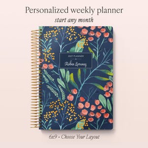 May include: A spiral-bound weekly planner with a dark blue cover featuring a botanical pattern of orange flowers and green leaves. A central rectangular label displays the text 2027 PLANNER and the name Robin Lorway. The planner measures 6x9 inches.