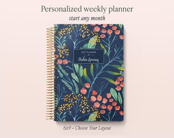 May include: A spiral-bound weekly planner with a dark blue cover featuring a botanical pattern of orange flowers and green leaves. A central rectangular label displays the text 2027 PLANNER and the name Robin Lorway. The planner measures 6x9 inches.