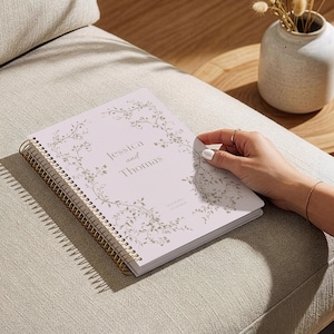 May include: A light pink spiral-bound planner with gold accents, featuring the names "Jessica and Thomas" and floral design. The planner is on a beige upholstered surface, with a small vase and wooden table in the background.