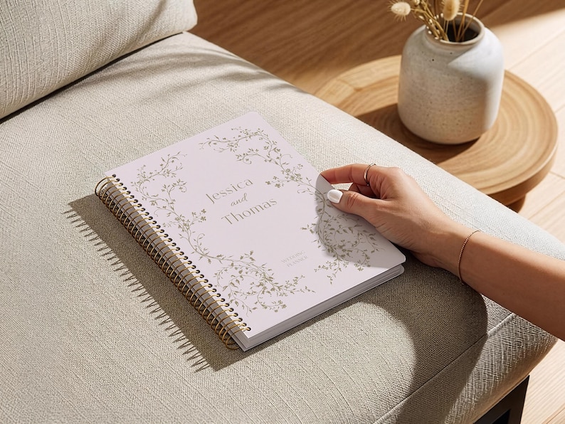 May include: A light pink spiral-bound planner with gold accents, featuring the names "Jessica and Thomas" and floral design. The planner is on a beige upholstered surface, with a small vase and wooden table in the background.