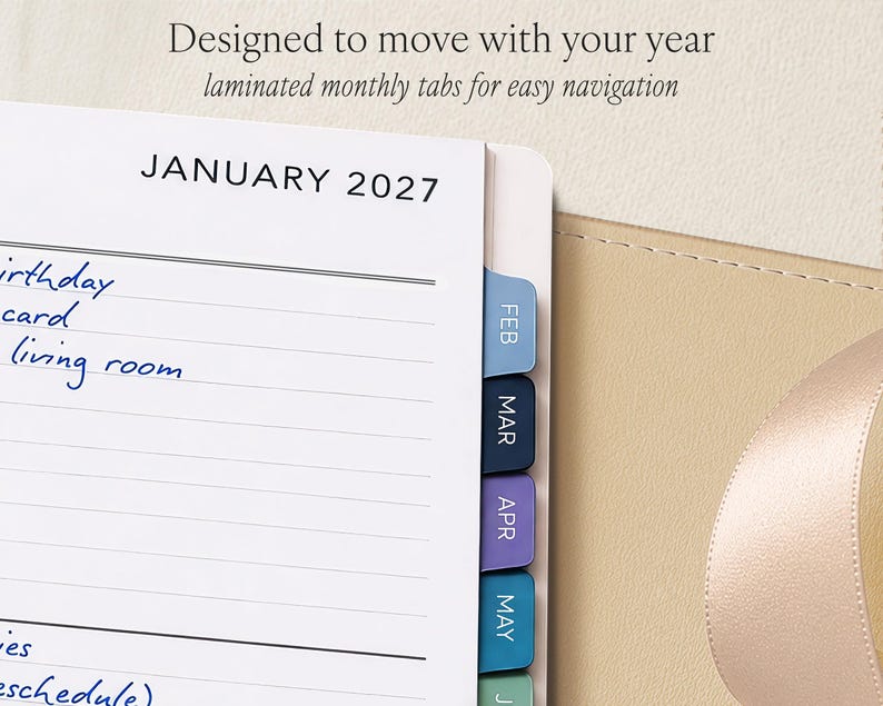May include: A planner page showing January 2027 with lined writing space and blue handwritten notes. The page features colorful laminated monthly tabs labeled FEB, MAR, APR, MAY, and JUN. Text at the top reads: Designed to move with your year.