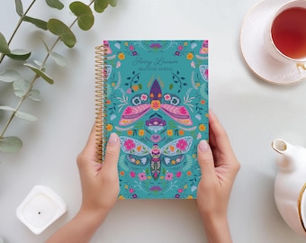 May include: A teal spiral-bound journal with a floral and moth design. The cover features the text "Avery Lawson GRATITUDE JOURNAL". The journal is held by two hands, with a cup of tea and a teapot in the background.