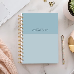 Peut inclure: Un agenda bleu 2025 avec le nom "CONNOR BAILY" imprimé sur la couverture. L'agenda est sur une surface en marbre blanc avec un ordinateur portable, une plante succulente en pot, un stylo et des écouteurs.