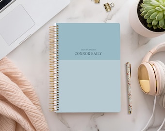 May include: A blue 2025 planner with the name "CONNOR BAILY" printed on the cover. The planner is on a white marble surface with a laptop, a potted succulent, a pen, and headphones.