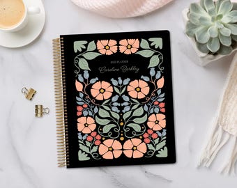 May include: A black 2023 planner with a gold spiral binding. The cover features a floral design in peach, green, and blue. The text "2023 Planner Caroline Berkley" is displayed on the cover.