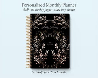 May include: A black 6x9 inch personalized monthly planner with gold spiral binding. The cover features a floral design in gold and pink, with the name "Ashley Murray" and the year "2027" printed on it. The planner has no weekly pages.