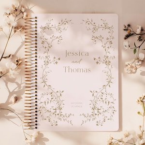 May include: A wedding planner with a spiral binding, featuring the names "Jessica and Thomas" and the words "Wedding Planner". The cover has a floral design in light green. White flowers are arranged around the planner.