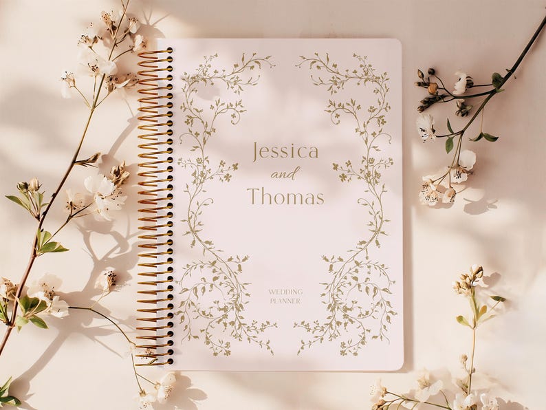 May include: A spiral-bound wedding planner with the names "Jessica and Thomas" and the words "Wedding Planner" printed on the cover. The cover has a floral design in a light green color. The planner is surrounded by white flowers.