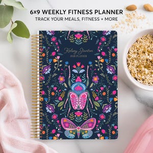 May include: A 6x9 inch weekly fitness planner with a navy blue cover featuring a floral and insect design. The planner has gold spiral binding and includes the text "Track Your Meals, Fitness + More". A bowl of oats and a pink water bottle are also visible.