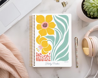 May include: A spiral-bound planner with a floral design featuring yellow flowers and teal leaves. The cover includes the text "Haley Parker" and "2023-28". A pen, headphones, and a succulent plant are also visible.